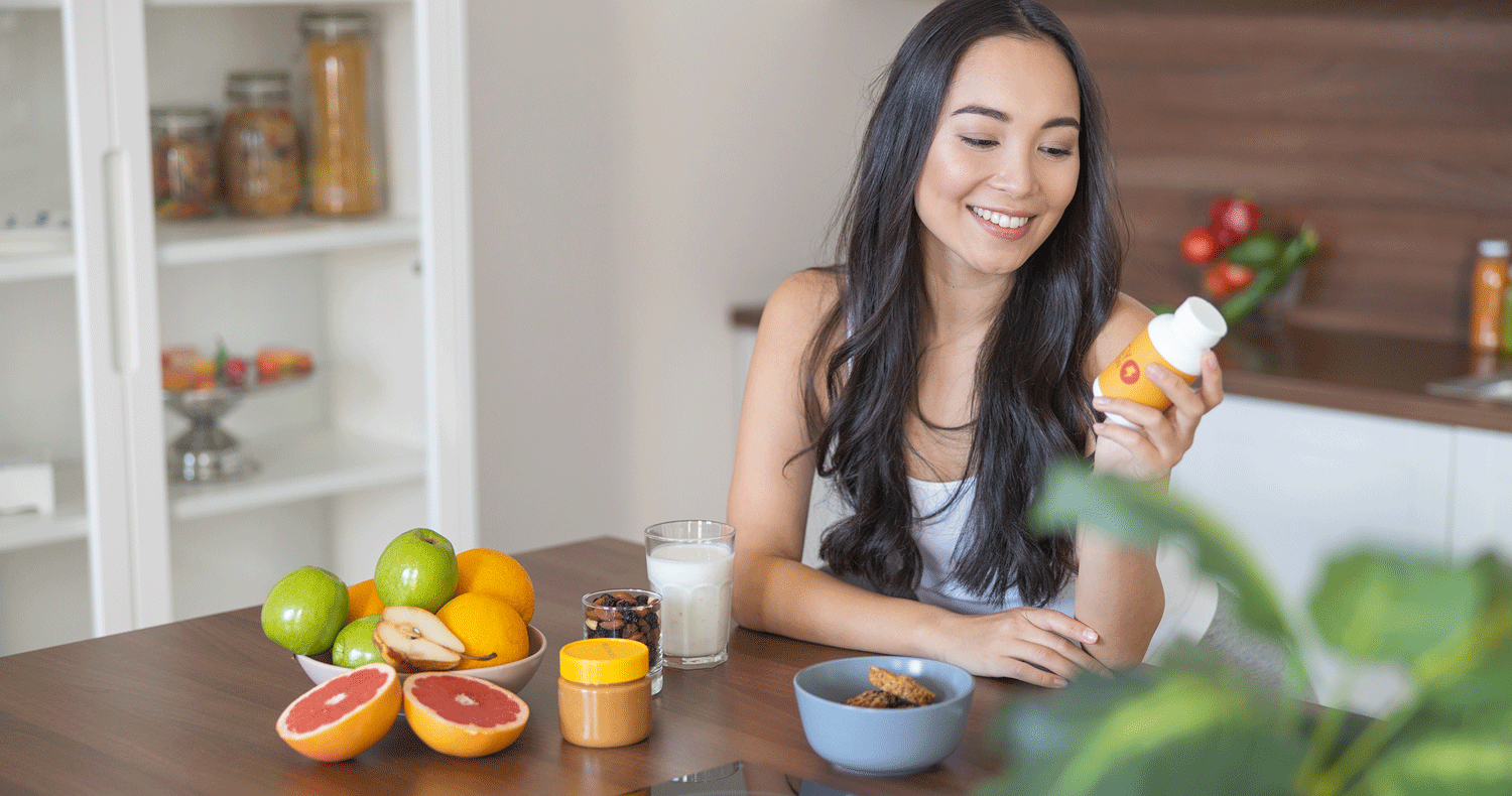Vegane-Nahrungsergänzungsmittel Junge Frau am Tisch hält ein Nahrungsergänzungsmittel in der Hand. Vor ihr frisches Obst