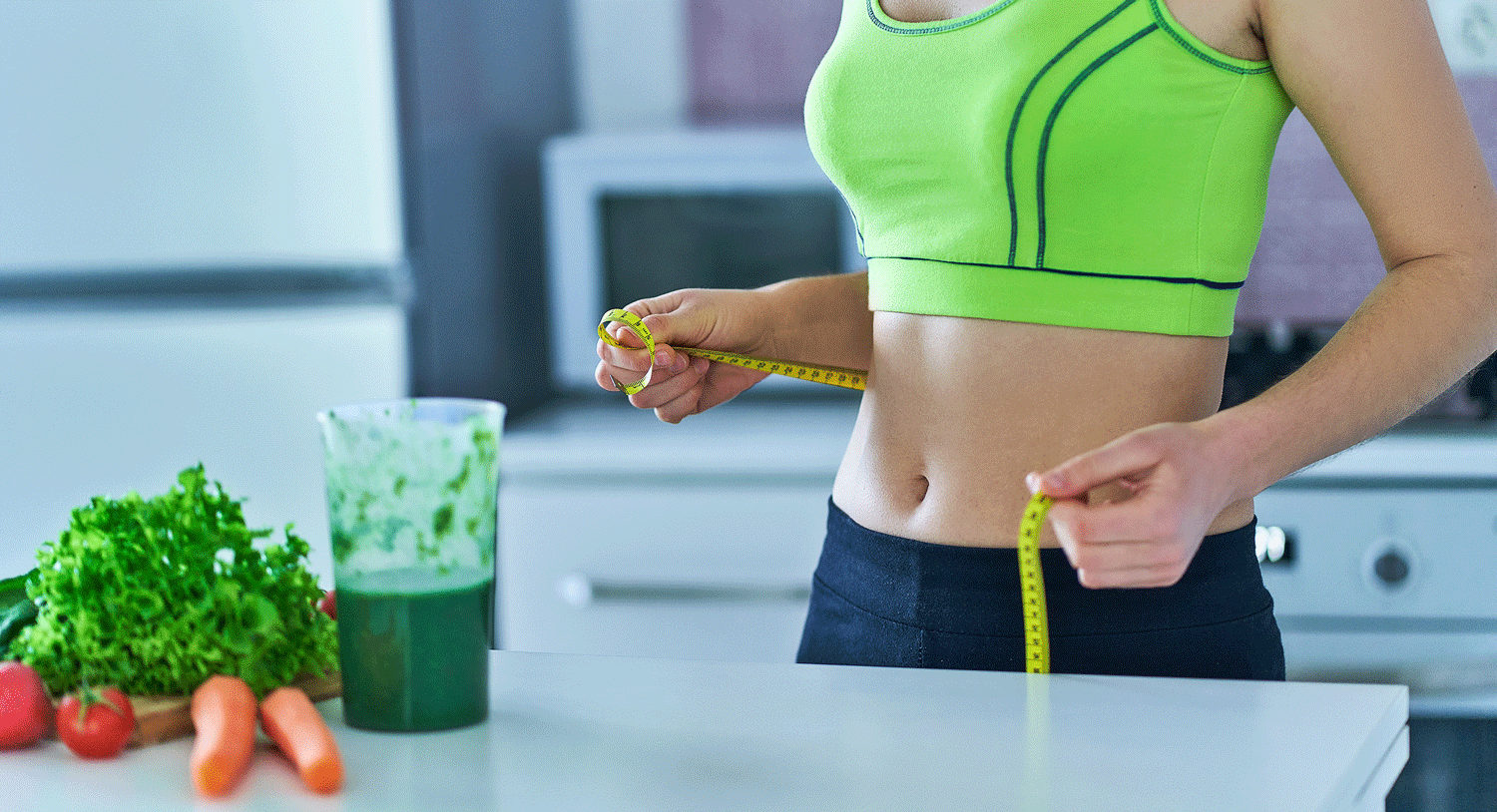 Nahrungsergänzungsmittel-zum-Abnehmen Junge Frau hält in der Küche ein Maßband um ihre Taille, vor Ihre ein grüner Smoothie und Gemüse